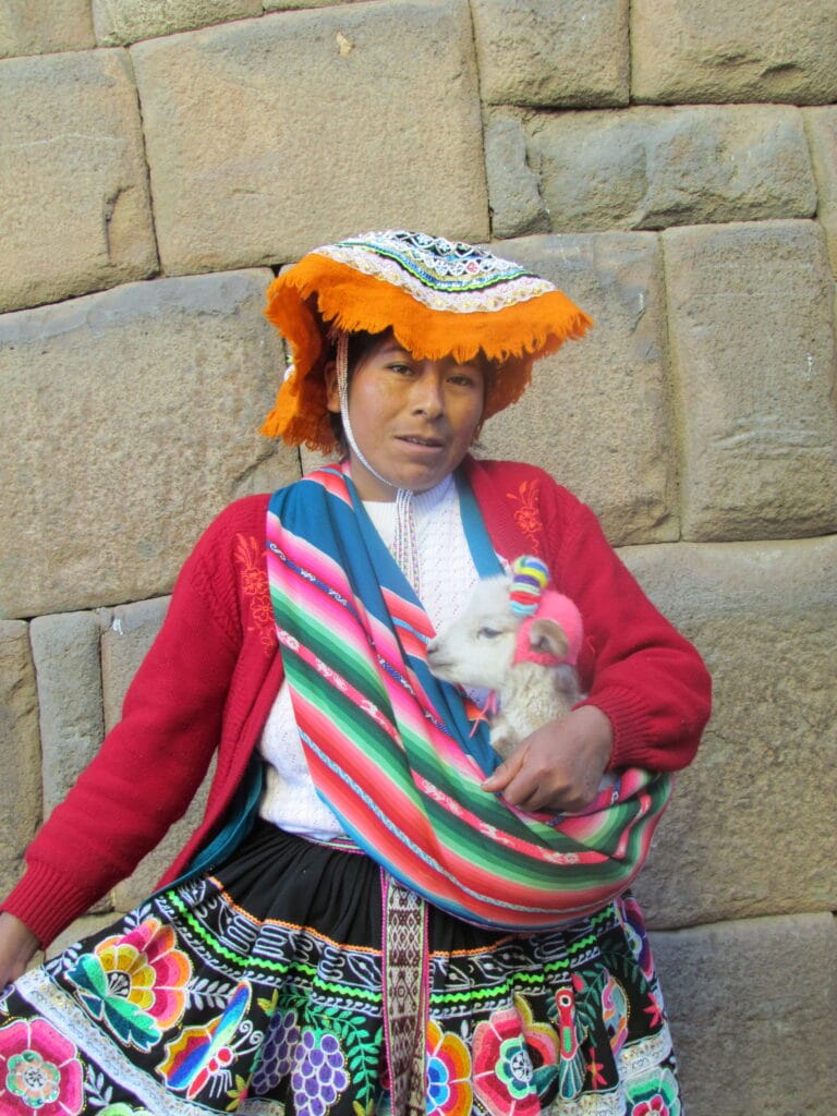 Woman in traditional Andean dress holding a lamb in a woven sling, stone wall behind, Cusco.